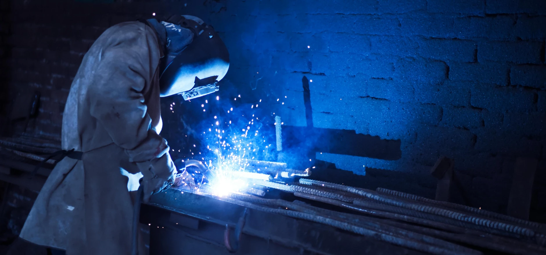 Industrial worker welding metal indoors, emitting bright blue sparks and smoke.