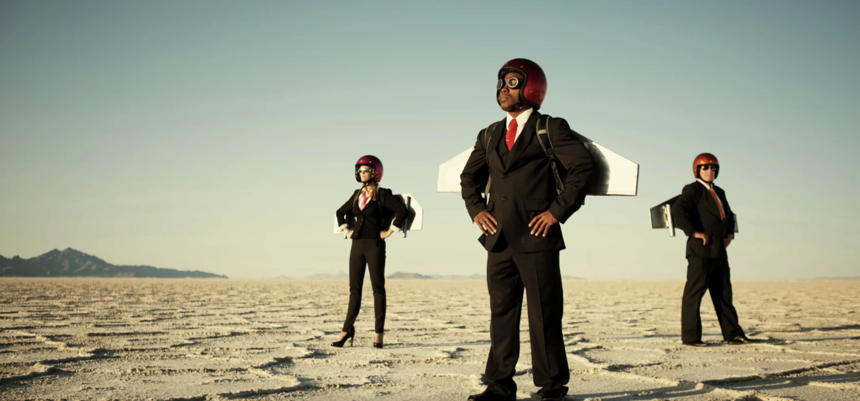 People in suits wearing rocket wings and helmets stand hands on hips looking out in the desert 