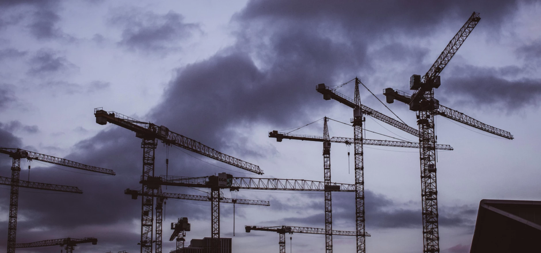 Silhouetted tower cranes against a moody twilight sky at a construction site.