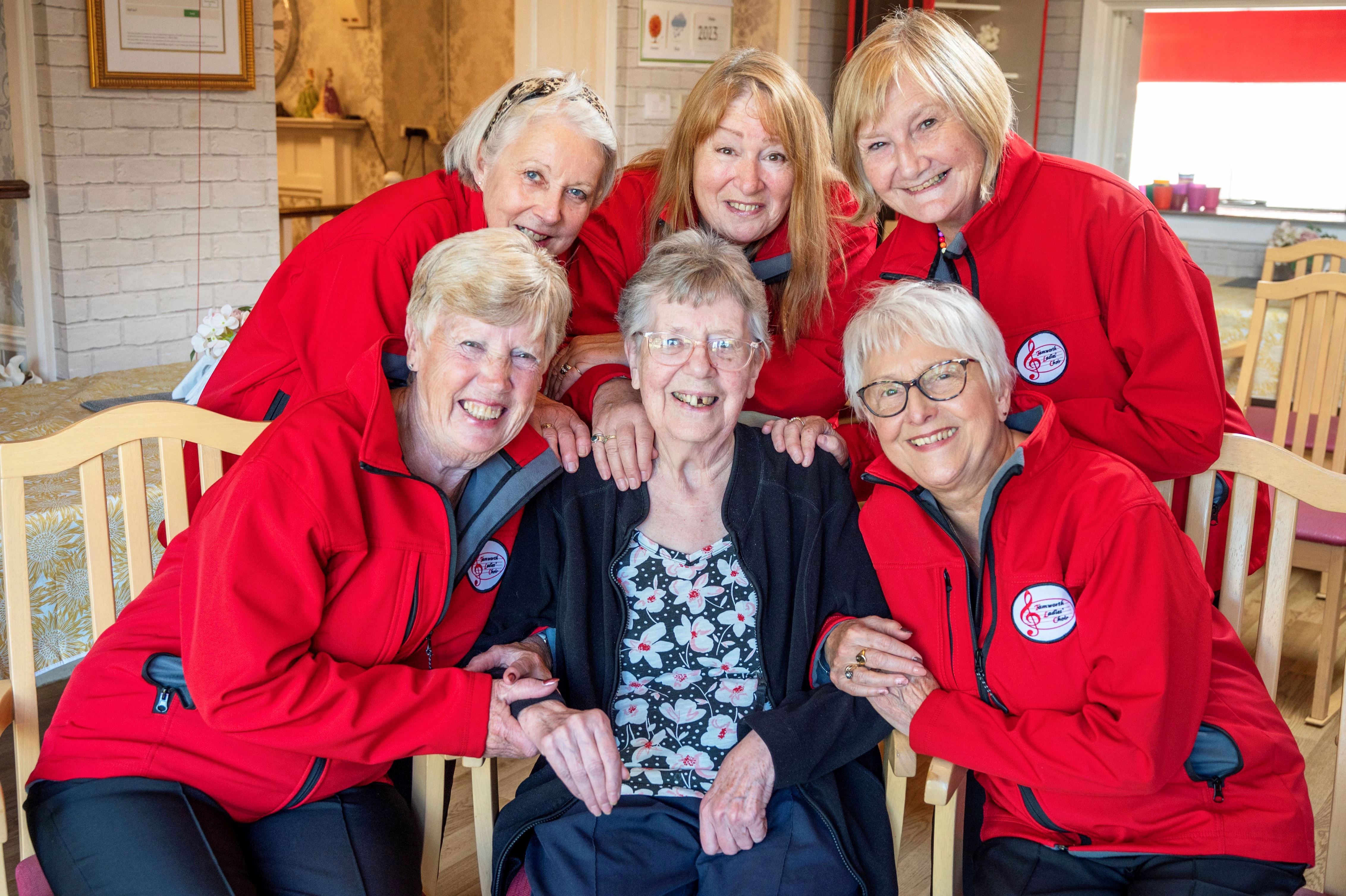 Ladies from Tamworth's Ladies choir in red fleeces, standing around care home resident and former member, Thelma Peet.