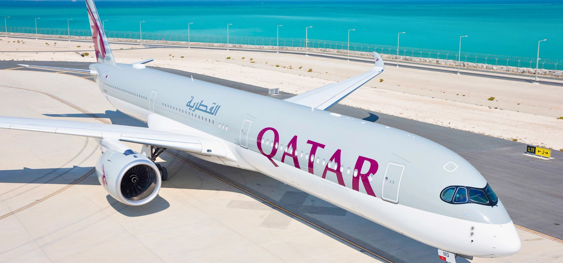 A Qatar Airways aircraft sits on a runway in front of the ocean.