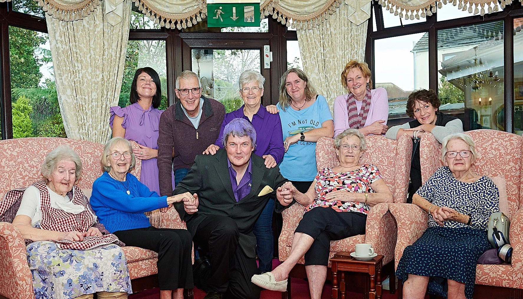 Activities coordinator Paul Hipwood with purple hair, the centre of a group of residents, family members and volunteers from the Alz Cafe. Everyone is smiling to the camera.