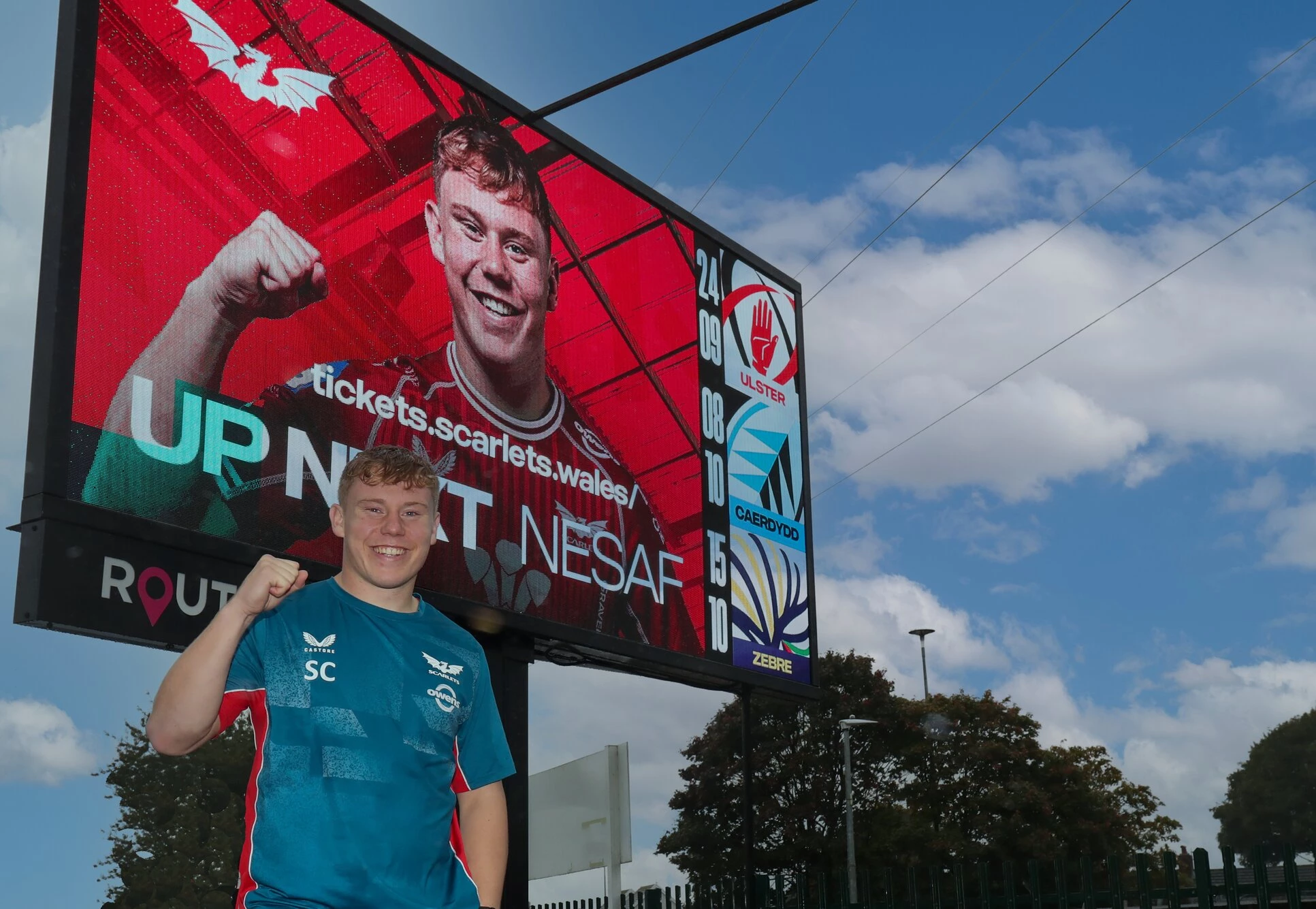 Scarlets fly half Sam Costelow with one of the Route Media digital signs