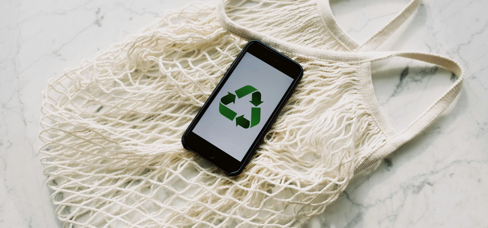 Overhead of smartphone with simple recycling sign on screen placed on white eco friendly mesh bag on marble table in room